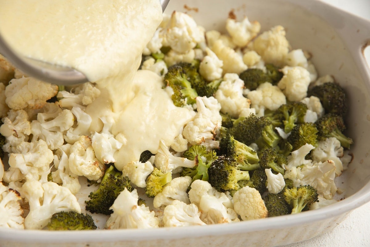 Pouring the creamy sauce into the baking dish with the roasted broccoli and cauliflower.