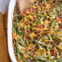 Large casserole dish of cheesy green bean casserole with chopped cooked bacon sprinkled on top and a napkin off to the side.