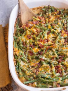 Large casserole dish of cheesy green bean casserole with chopped cooked bacon sprinkled on top and a napkin off to the side.