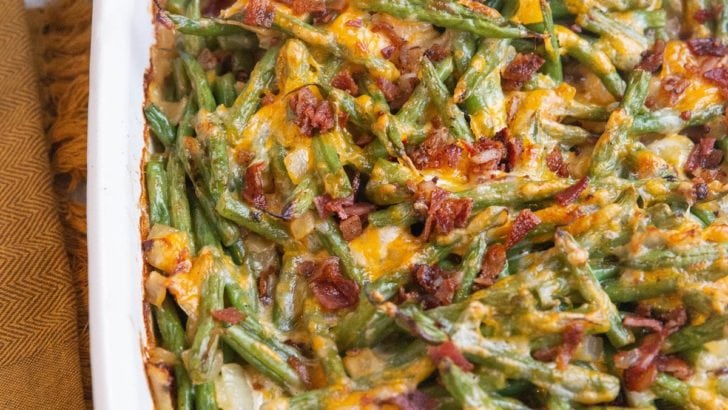Large casserole dish of cheesy green bean casserole with chopped cooked bacon sprinkled on top and a napkin off to the side.