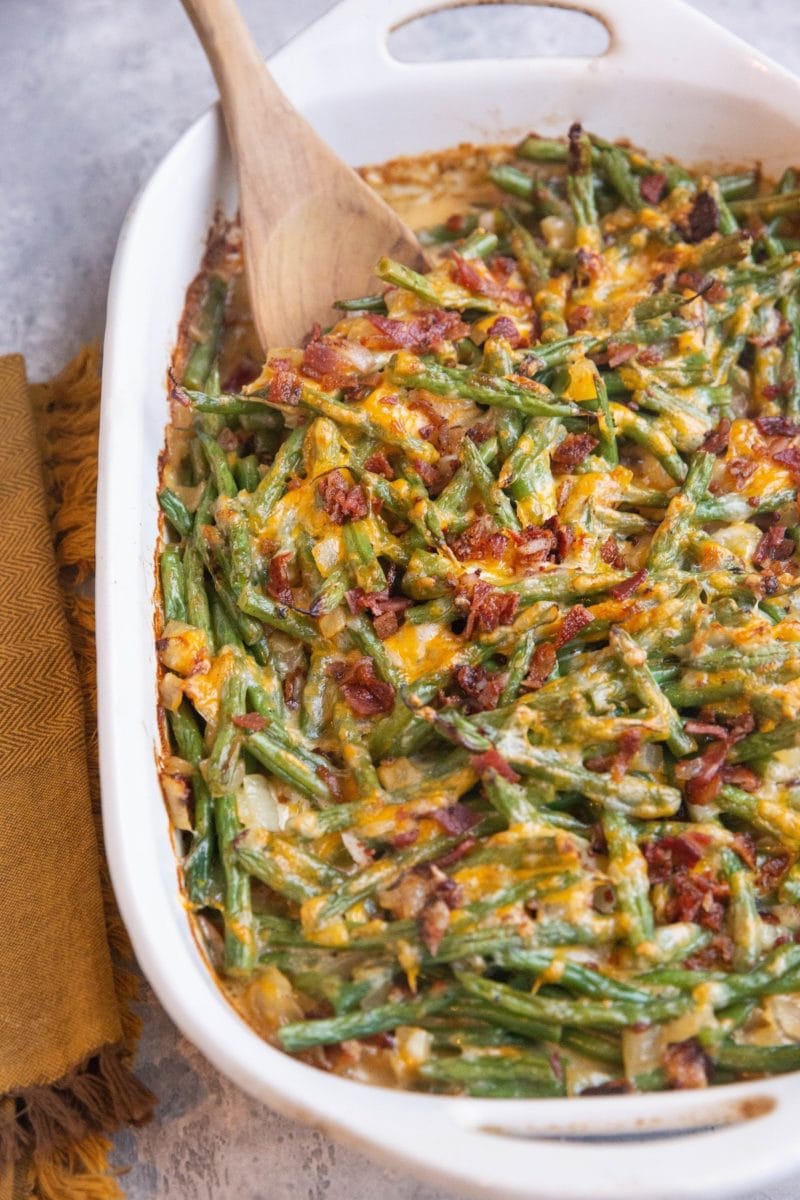 Large casserole dish of cheesy green bean casserole with chopped cooked bacon sprinkled on top and a napkin off to the side.