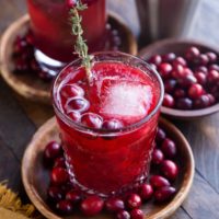 Two cranberry cocktails on wooden plates with fresh cranberries all around and a cocktail shaker in the background.