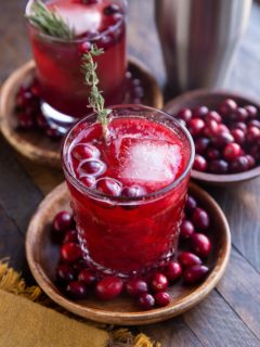 Two cranberry cocktails on wooden plates with fresh cranberries all around and a cocktail shaker in the background.