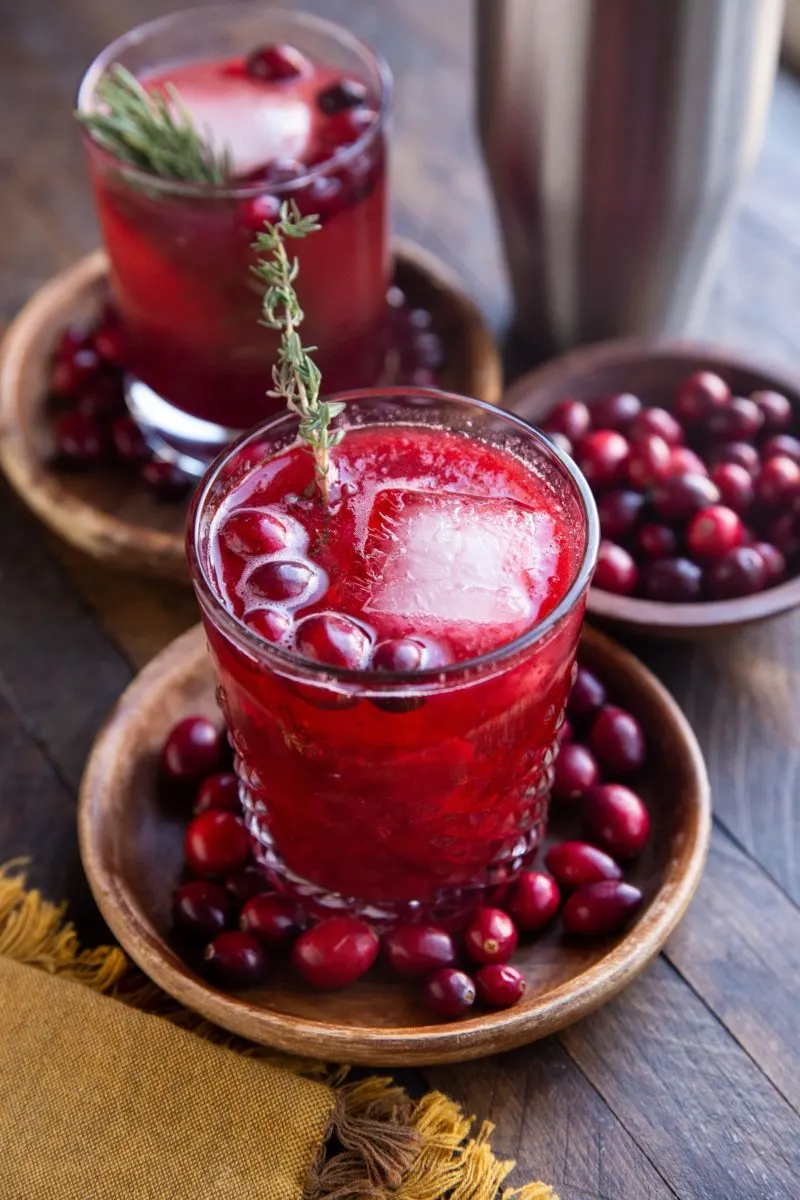 Two cranberry cocktails on wooden plates with fresh cranberries all around and a cocktail shaker in the background.