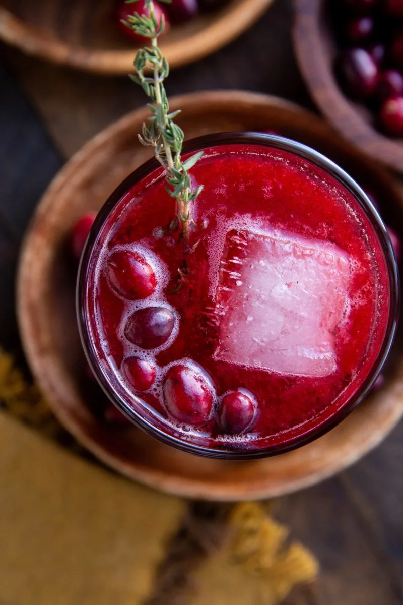 Close up top down image of a cranberry cocktail on a dark wooden backdrop with a sprig of fresh thyme in it.