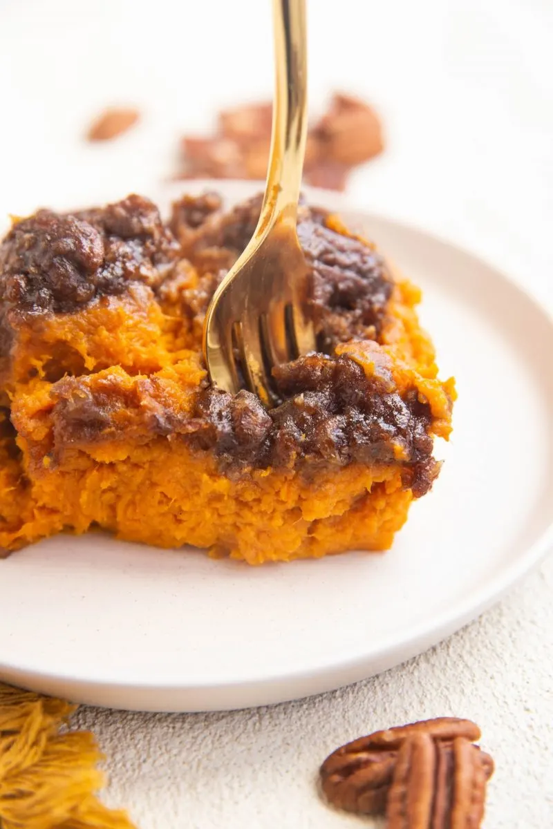 Slice of sweet potato casserole on a white plate with a gold fork taking a bite out.