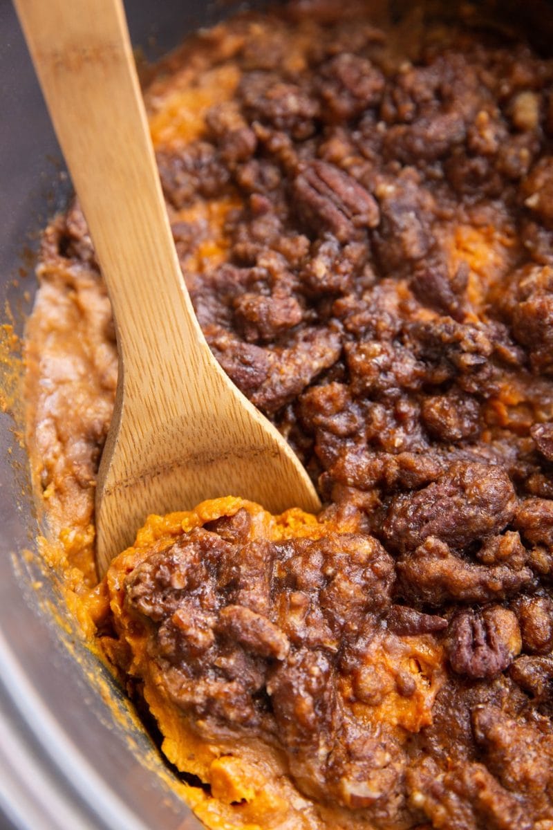 Fully cooked sweet potato casserole with pecan topping inside of a crock pot with a wooden spoon taking a scoop out.