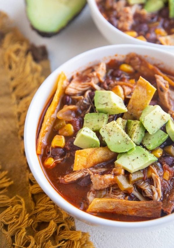 Two white bowls of chicken tortilla soup with avocado on top and a golden napkin to the side.