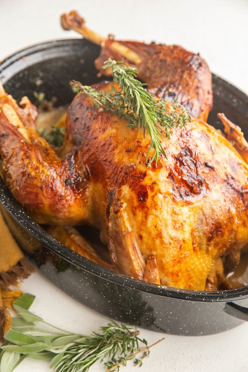 Roast turkey in a roasting pan with fresh herbs on top and fresh herbs to the side.