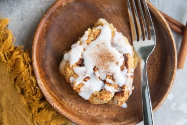 Horizontal photo of a cinnamon roll on a wooden plate with a fork and a napkin to either side.