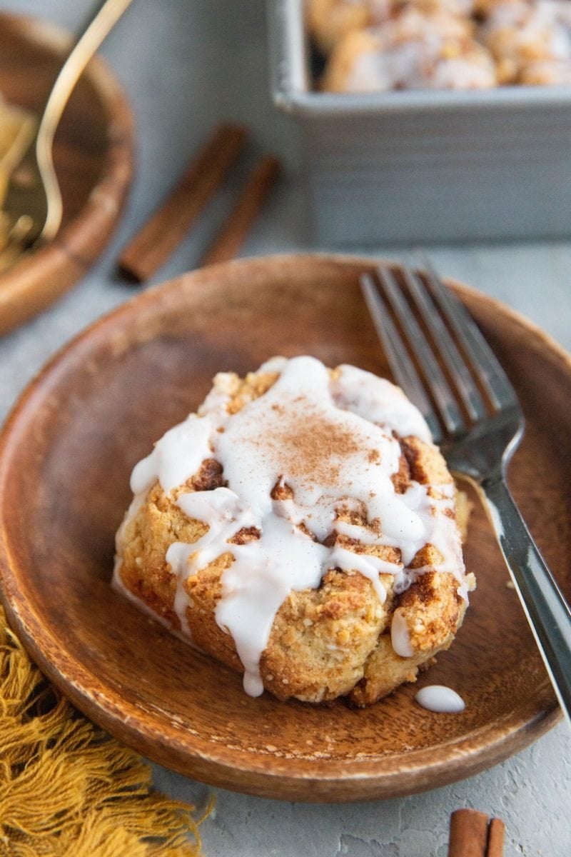 Cinnamon roll on a wooden plate with glaze on top and tray of cinnamon rolls in the background.