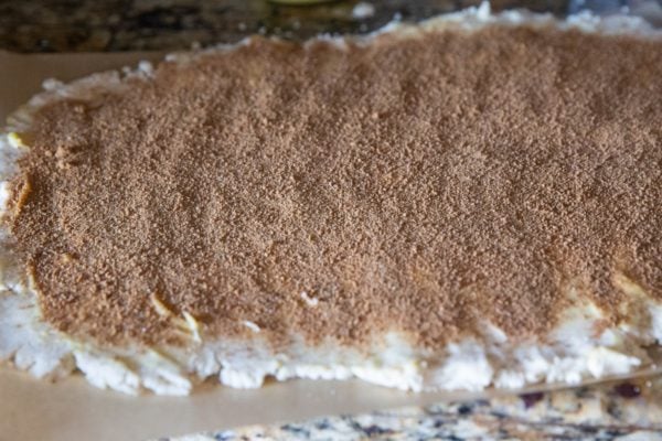 Flat cinnamon roll dough on parchment paper, sprinkled with cinnamon and sugar, ready to be rolled.