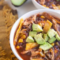 Two white bowls of chicken tortilla soup with avocado on top and a golden napkin to the side.