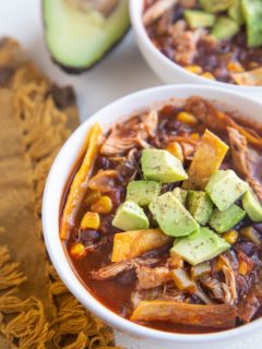 Two white bowls of chicken tortilla soup with avocado on top and a golden napkin to the side.