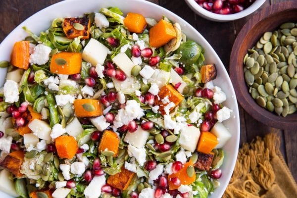 Horizontal top down photo of white bowl of brussel sprout salad with a bowl of pumpkin seeds and pomegranate seeds to the side.