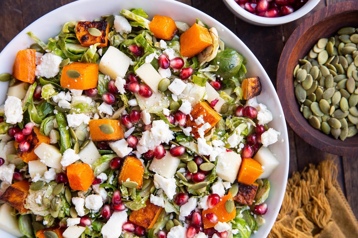 Horizontal top down photo of white bowl of brussel sprout salad with a bowl of pumpkin seeds and pomegranate seeds to the side.