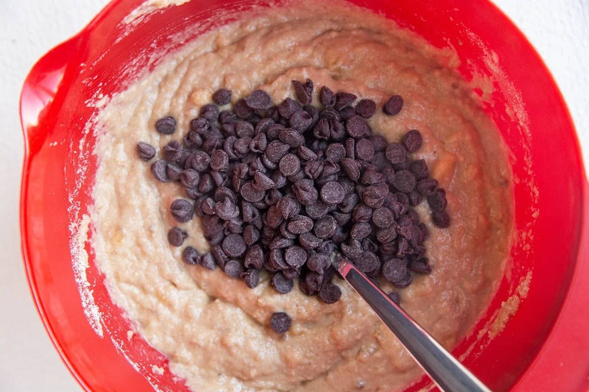 Banana muffin batter in a red mixing bowl with chocolate chips on top, ready to be mixed in.