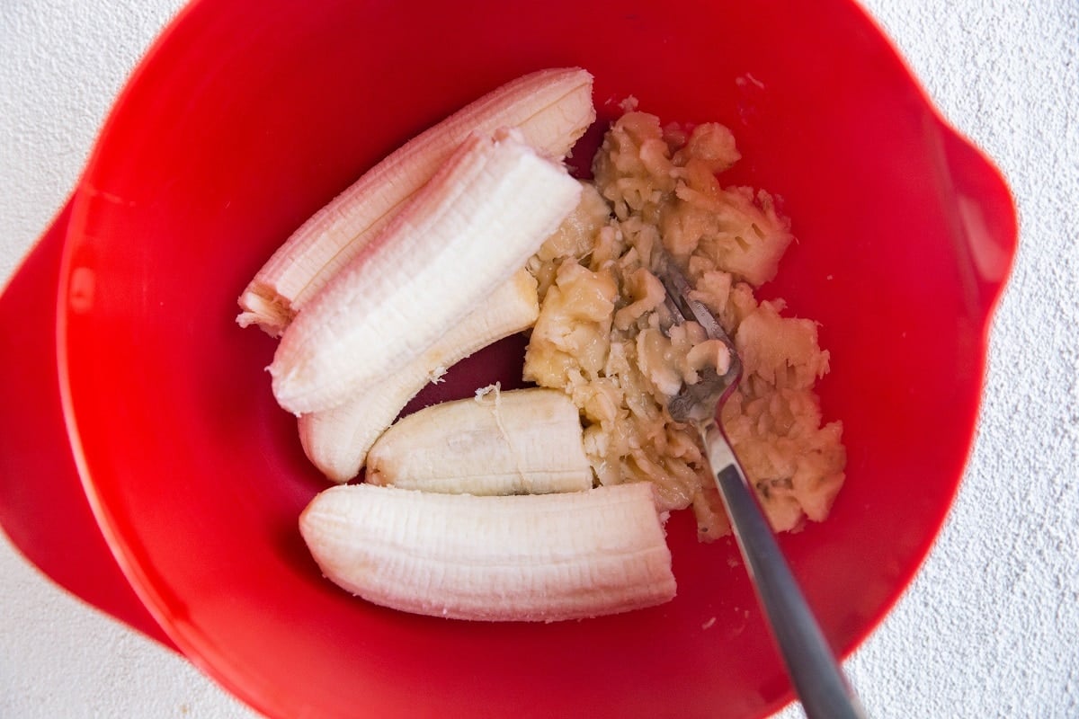 Bananas being mashed by a fork in a red mixing bowl.