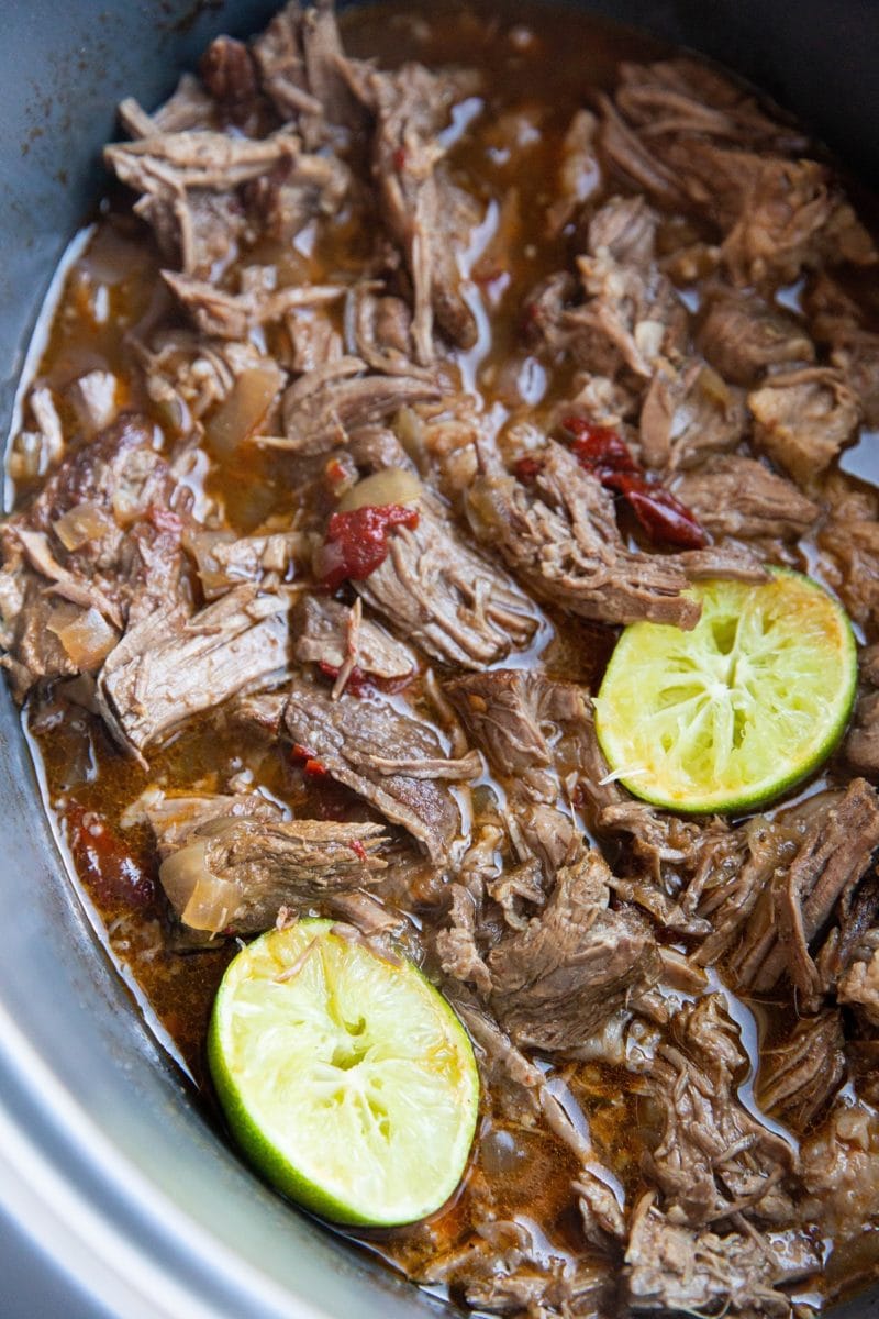 Close up image of a slow cooker full of shredded barbacoa beef with juiced limes inside.