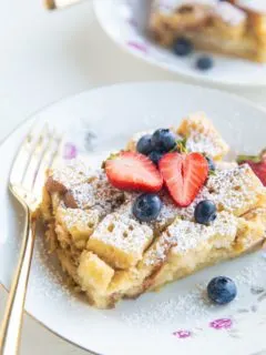 Slices of bread pudding in china bowls with fresh berries on top and gold forks to the side.