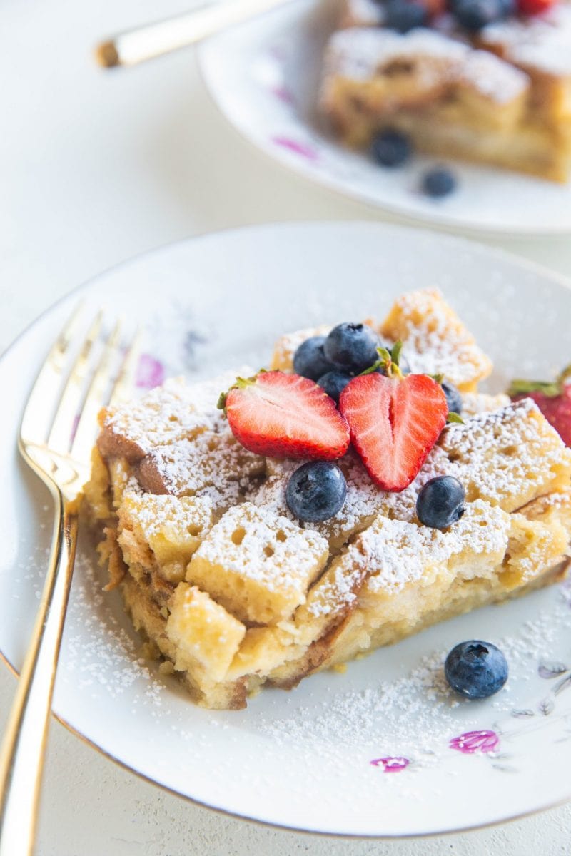 Slices of bread pudding in china bowls with fresh berries on top and gold forks to the side.