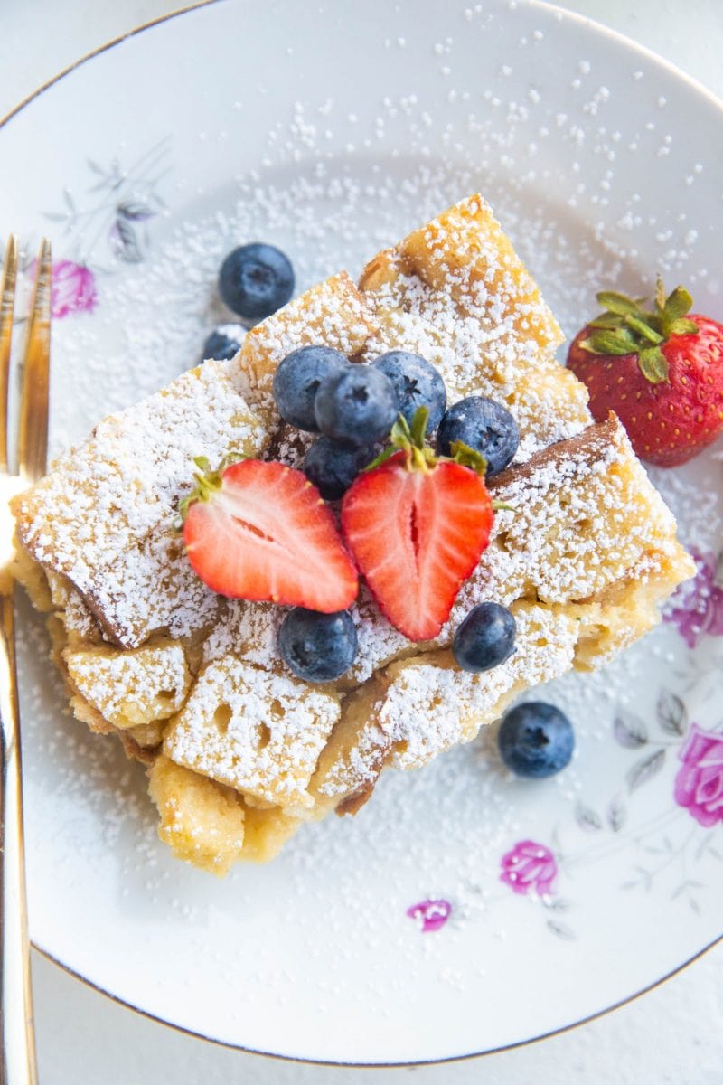 Top down photo of a slice of bread pudding sprinkled with powdered sugar in a pretty bowl with fresh strawberries and blueberries on top.