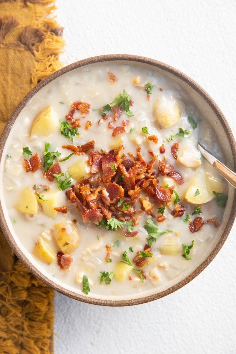 Top down photo of a ceramic bowl of clam chowder with cooked bacon on top and a golden napkin to the side.
