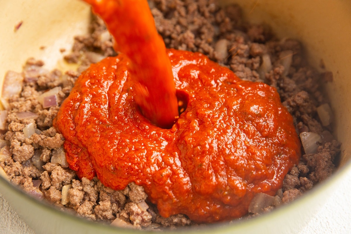 Pouring tomato sauce into a pot with ground beef and onions.