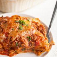 White plate of a large slice of gluten-free lasagna with the casserole dish of lasagna in the background.