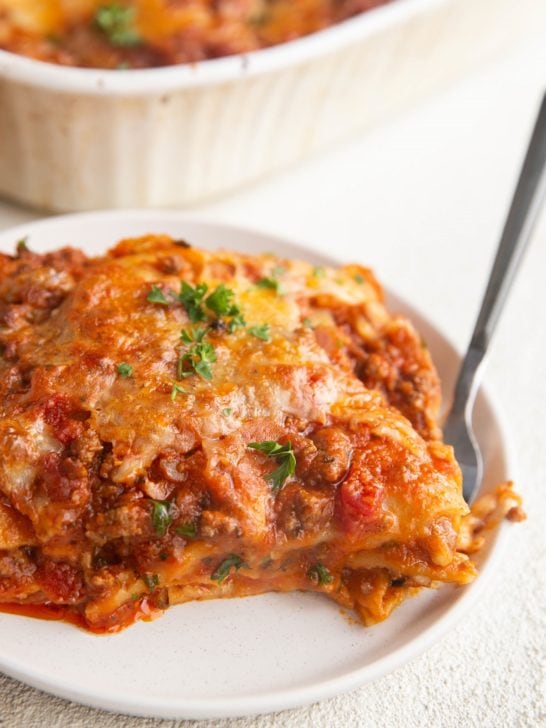 White plate of a large slice of gluten-free lasagna with the casserole dish of lasagna in the background.