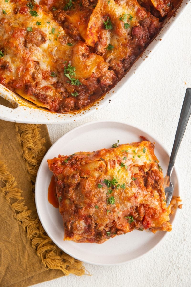 Top down image of a white plate of lasagna and a casserole dish, ready to be eaten.