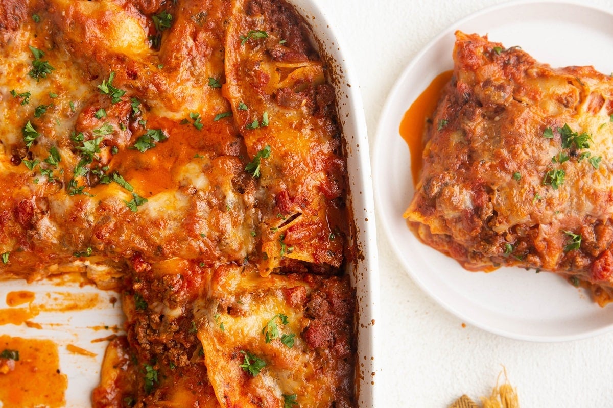 Horizontal photo of a white plate with a slice of lasagna next to the casserole dish full of lasagna.