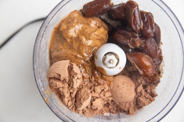 Ground oats, dates, almond butter, and cocoa powder in a food processor, ready to be blended.