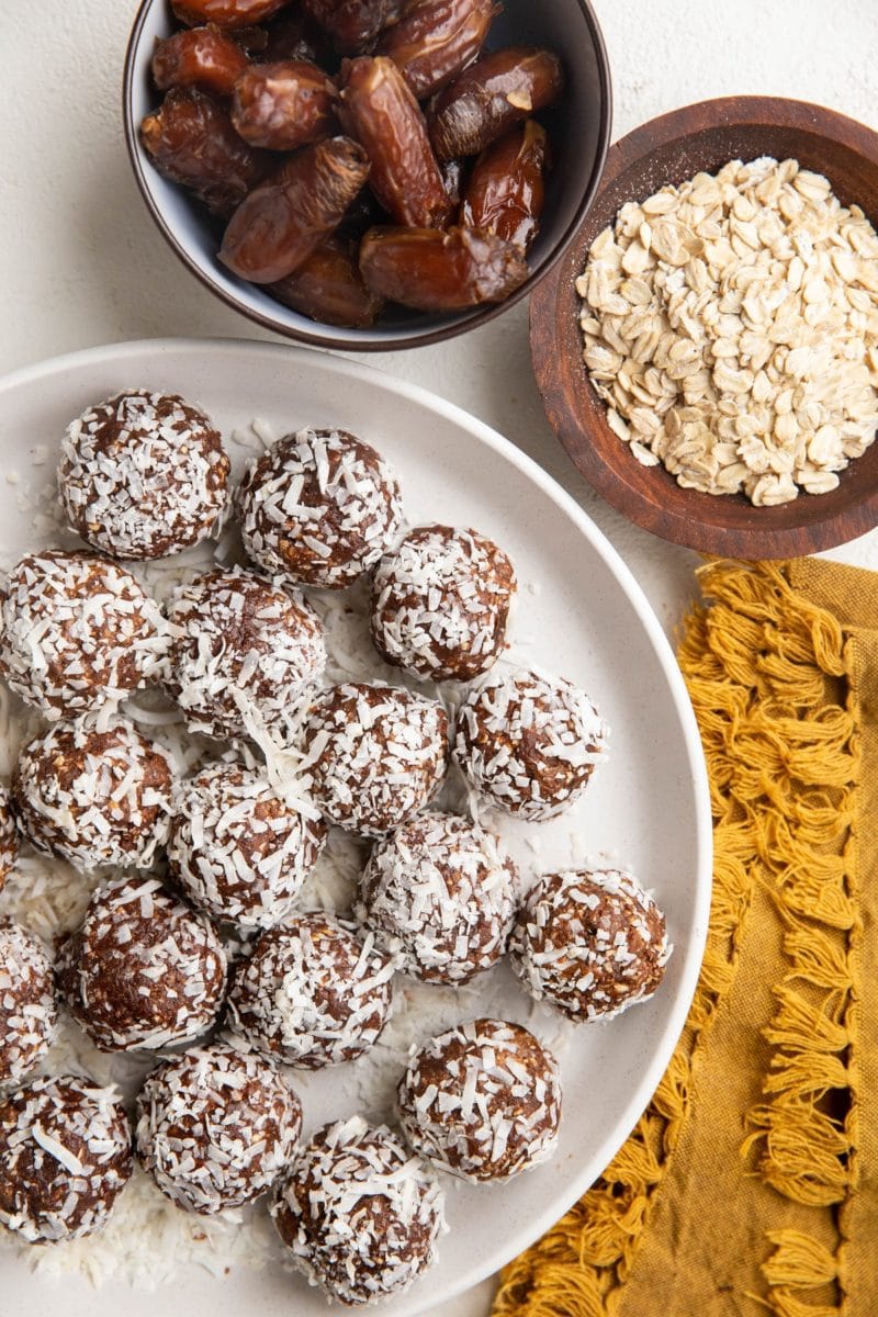 White plate of chocolate peanut butter energy bites with a bowl of oats and a bowl of dates to the side.
