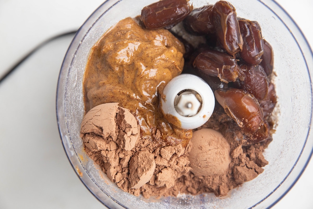 Ground oats, dates, almond butter, and cocoa powder in a food processor, ready to be blended.