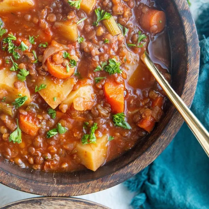 Two wooden bowls of Lentil soup with a blue napkin to the side and fresh chopped parsley to the side. A gold spoon inside of the bowl, ready to eat.