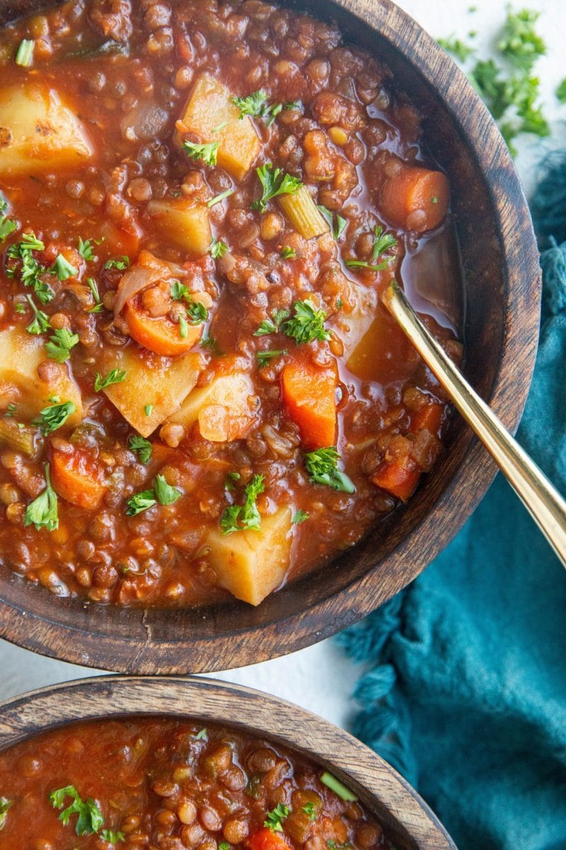 Two wooden bowls of Lentil soup with a blue napkin to the side and fresh chopped parsley to the side. A gold spoon inside of the bowl, ready to eat.