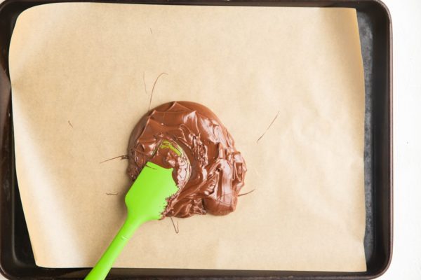 Melted chocolate on a sheet of parchment paper on a baking sheet with a rubber spatula, about to be spread.