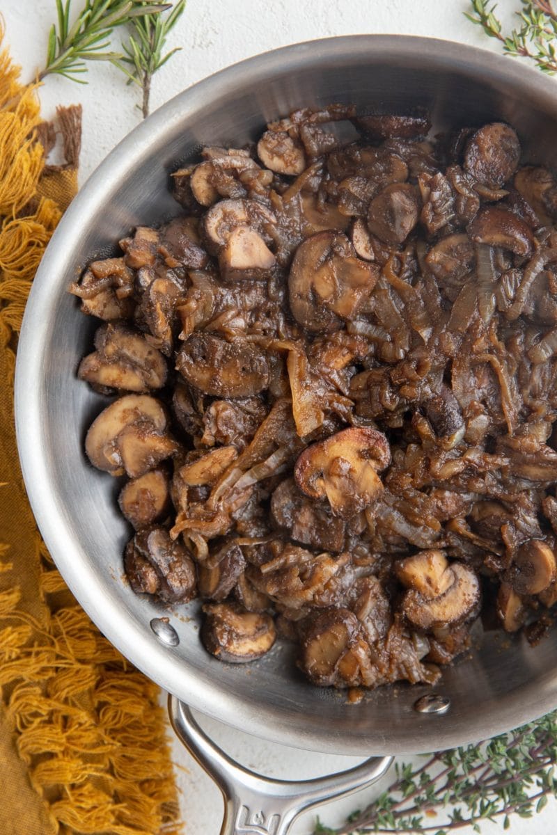 Stainless steel skillet with sauteed mushrooms and onions with fresh herbs to the side.