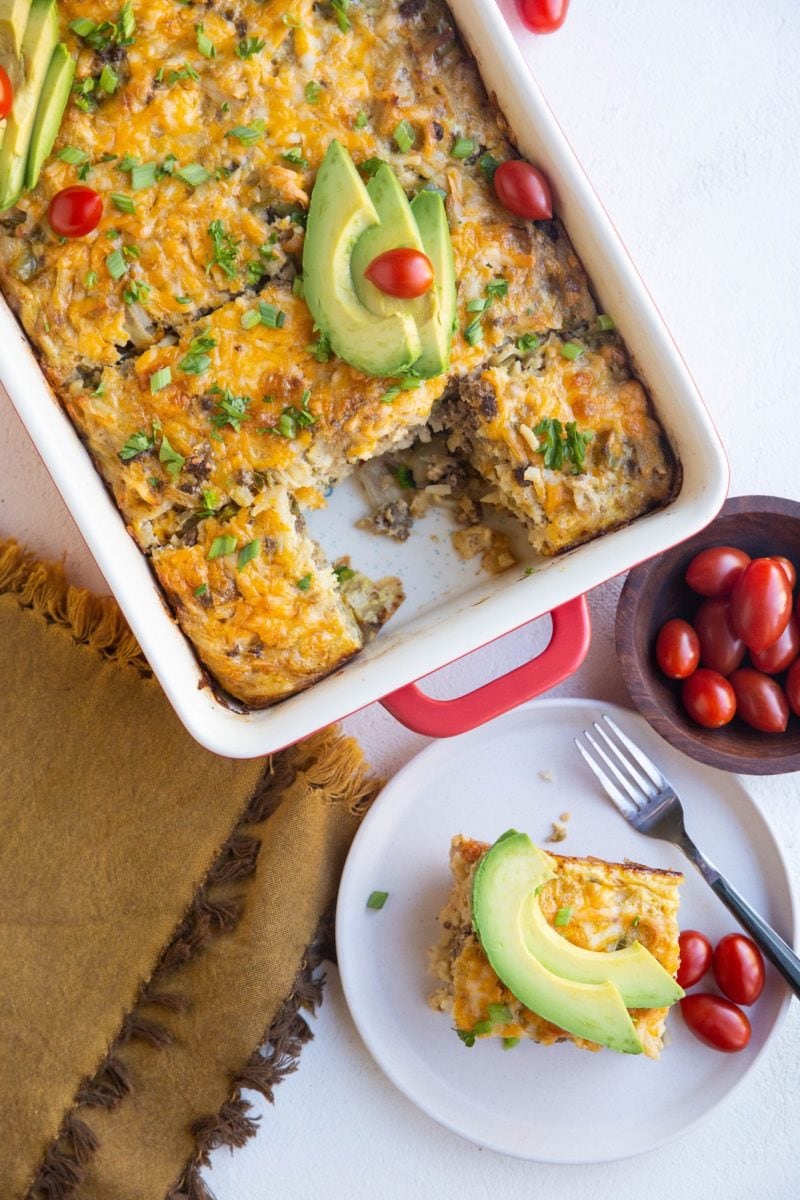 Casserole dish of taco breakfast casserole on a white background with a plate of casserole and cherry tomatoes to the side.