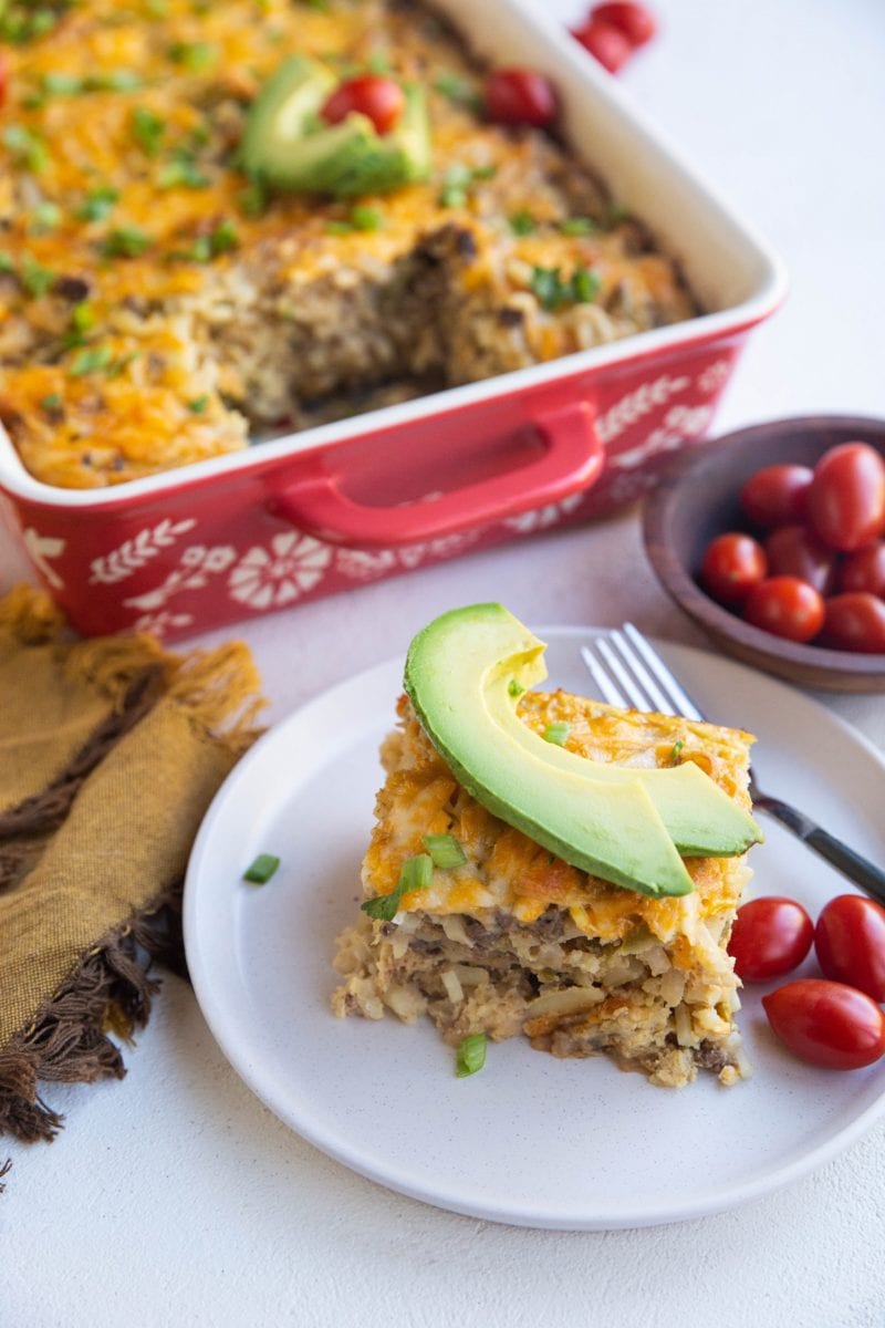 white plate with a hunk of breakfast casserole on it with slices of avocado on top with the casserole in the background.