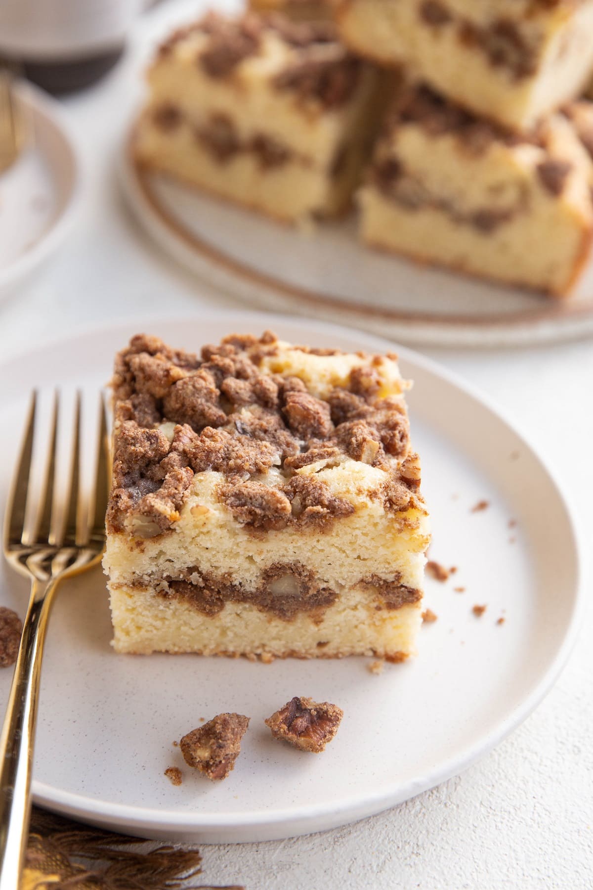 White plate with a single slice of coffee cake with a golden fork to the side and a plate of coffee cake slices in the background.