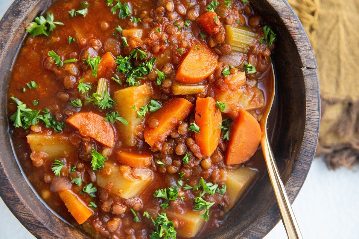 Horizontal image of a wooden bowl of lentil soup with a gold napkin to the side and a gold spoon in the bowl of soup.