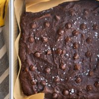 Square baking pan with healthy banana brownies sprinkled with sea salt and chocolate chips fresh out of the oven. A fresh banana and a napkin to the sides of the pan.