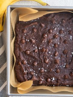 Square baking pan with healthy banana brownies sprinkled with sea salt and chocolate chips fresh out of the oven. A fresh banana and a napkin to the sides of the pan.