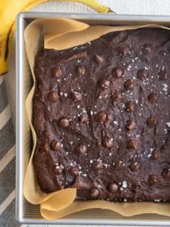 Square baking pan with healthy banana brownies sprinkled with sea salt and chocolate chips fresh out of the oven. A fresh banana and a napkin to the sides of the pan.