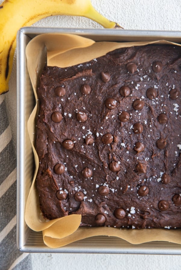Square baking pan with healthy banana brownies sprinkled with sea salt and chocolate chips fresh out of the oven. A fresh banana and a napkin to the sides of the pan.