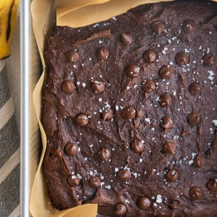 Square baking pan with healthy banana brownies sprinkled with sea salt and chocolate chips fresh out of the oven. A fresh banana and a napkin to the sides of the pan.