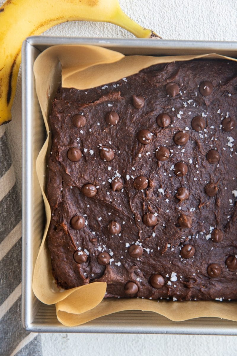 Square baking pan with healthy banana brownies sprinkled with sea salt and chocolate chips fresh out of the oven. A fresh banana and a napkin to the sides of the pan.