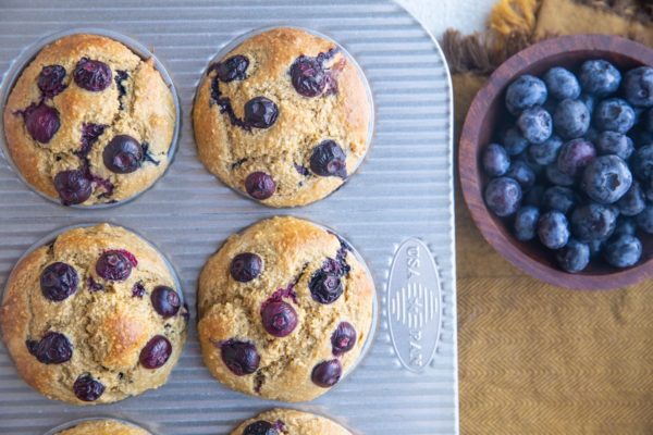 Blueberry Banana Oatmeal Muffins in a muffin tin, fresh out of the oven, with fresh blueberries in a wood bowl to the side.